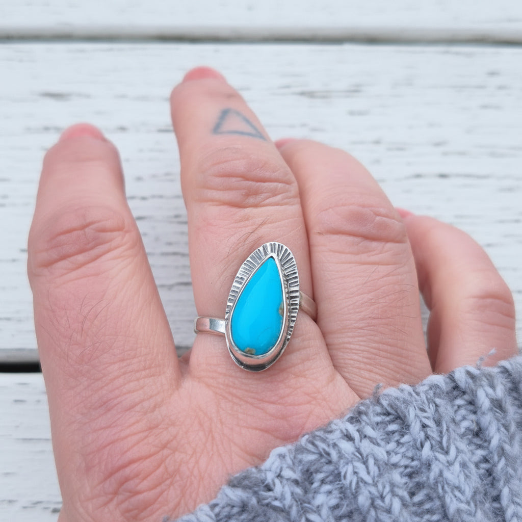 Hand wearing a silver ring with a blue stone against a blurred natural background